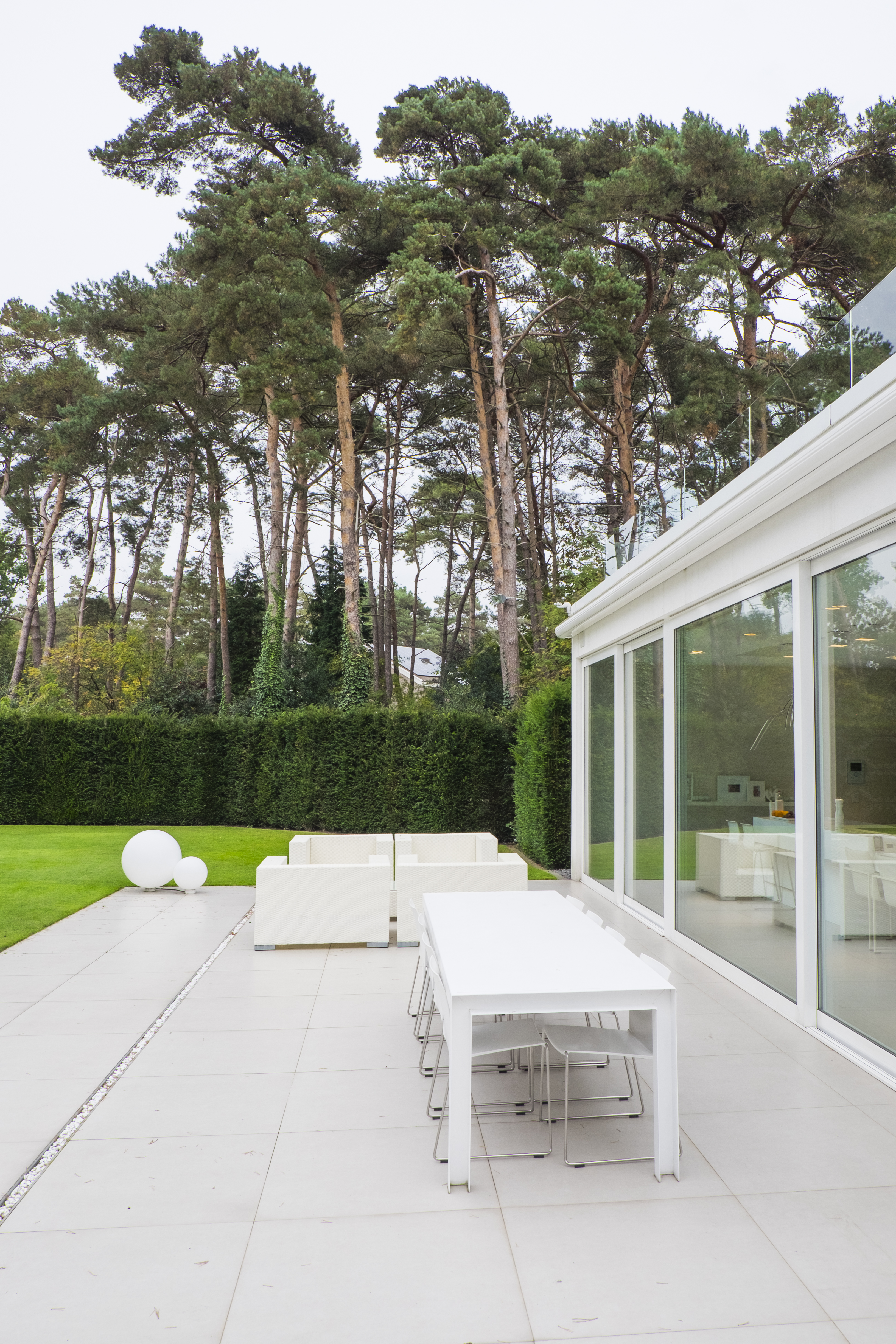 L'image montre une terrasse moderne avec de grandes portes coulissantes donnant sur un jardin luxuriant. Les fenêtres sont blanches, complétant le design minimaliste du mobilier extérieur. Les portes vitrées offrent une transition fluide entre l'intérieur et l'extérieur, renforçant l'impression d'espace et de légèreté.