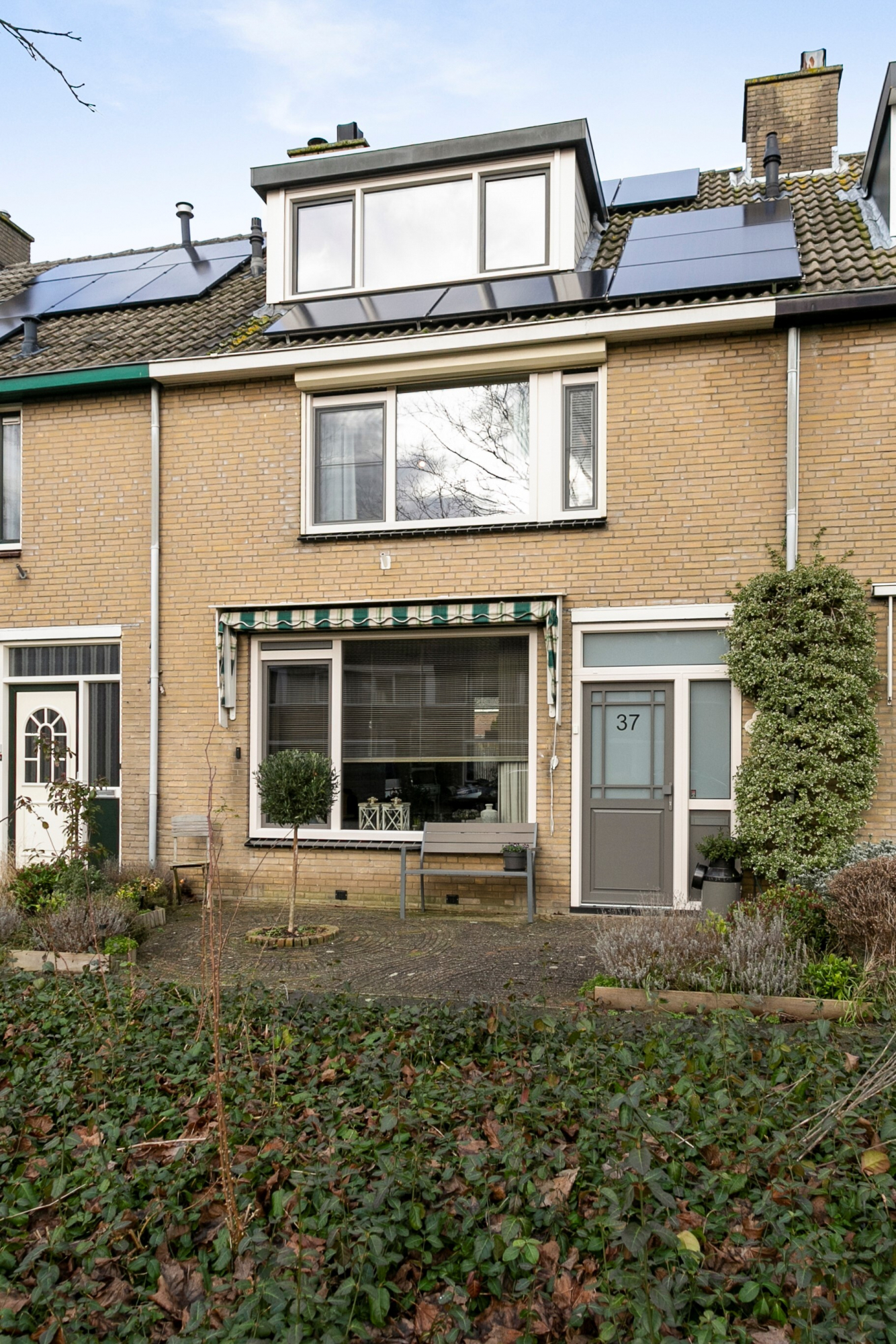 Het huis heeft een moderne gevel met grote kozijnen en een strakke deur. Op de begane grond is er een breed kozijn met een gestreepte zonwering, terwijl de bovenverdieping een rij kozijnen heeft die veel natuurlijk licht binnenlaten. De deur is eigentijds, met een matglazen paneel en een duidelijk zichtbaar huisnummer.