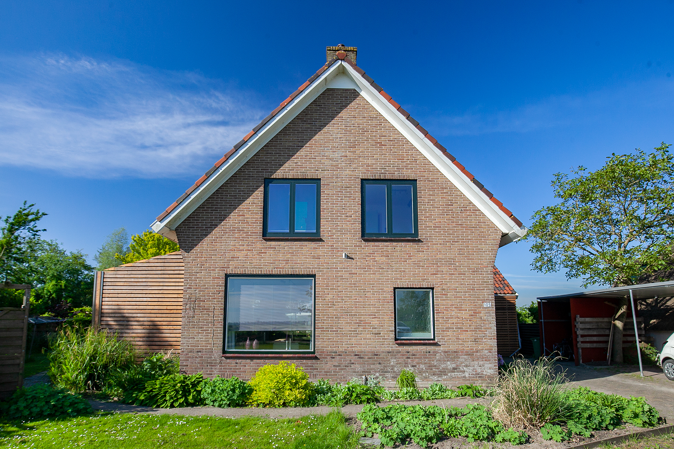Het huis heeft een traditionele bakstenen gevel met een zadeldak. Aan de voorkant zijn er drie grote kozijnen, twee op de bovenverdieping en één op de begane grond, die veel natuurlijk licht binnenlaten. De kozijnen zijn omlijst met donker materiaal, wat contrasteert met het lichte metselwerk. Er is geen zichtbare deur aan deze kant van het huis.