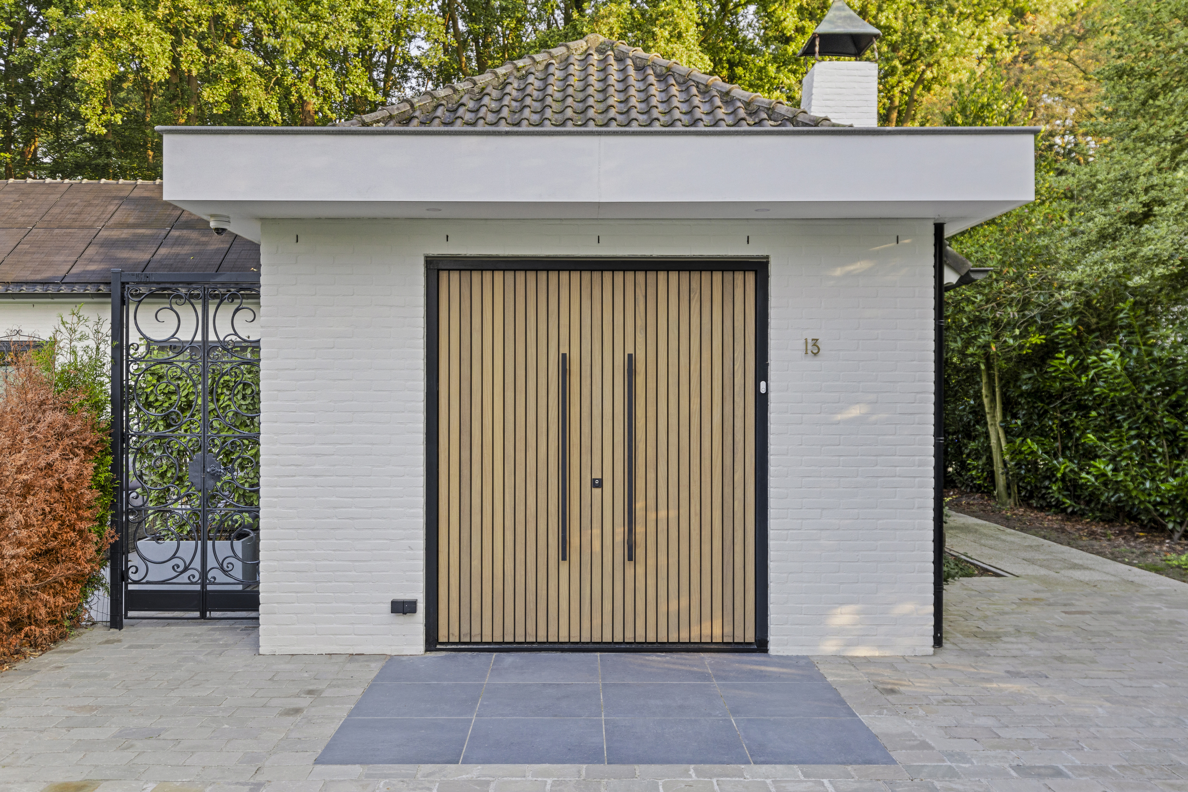 De afbeelding toont een moderne garagedeur met verticale houten latten, omlijst door een witte bakstenen muur. De deur wordt geflankeerd door een decoratief zwart smeedijzeren hek aan de linkerkant. De omgeving is rustig, met groen op de achtergrond.