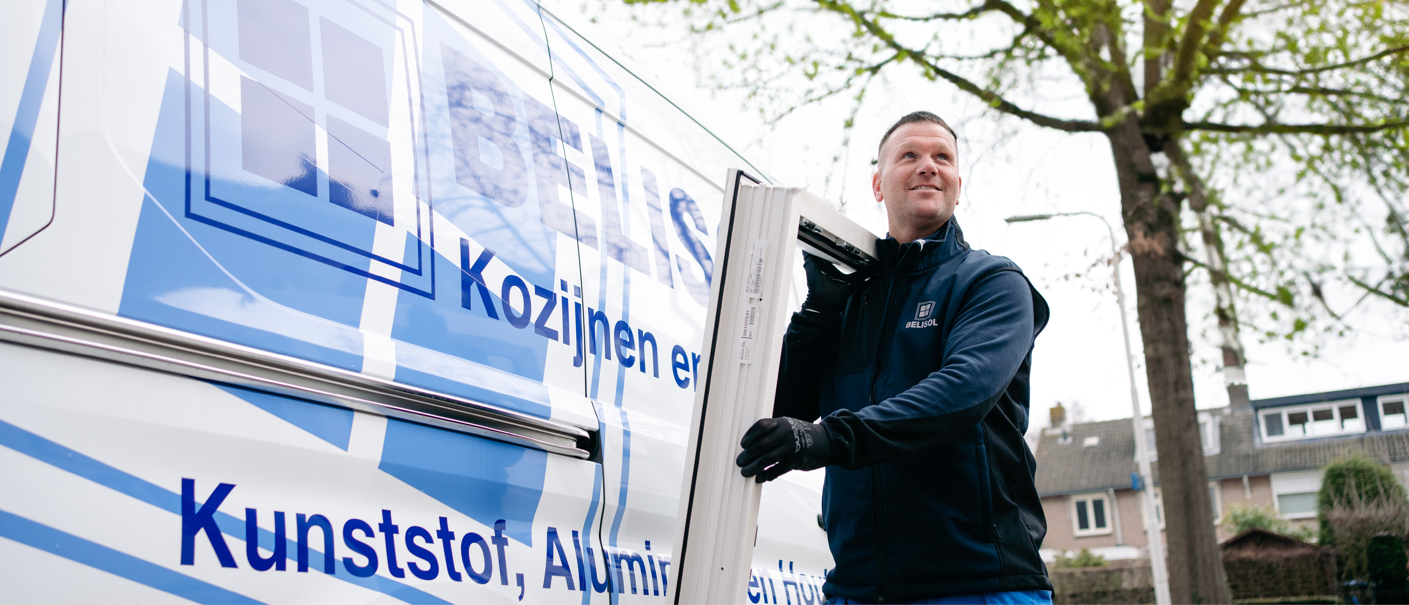 De afbeelding toont een man die een kozijn vasthoudt naast een busje. Het busje is voorzien van het logo en tekst van het bedrijf dat kozijnen en deuren aanbiedt. De man draagt een uniform met het bedrijfslogo, wat aangeeft dat hij waarschijnlijk een medewerker is die betrokken is bij installatie of levering.