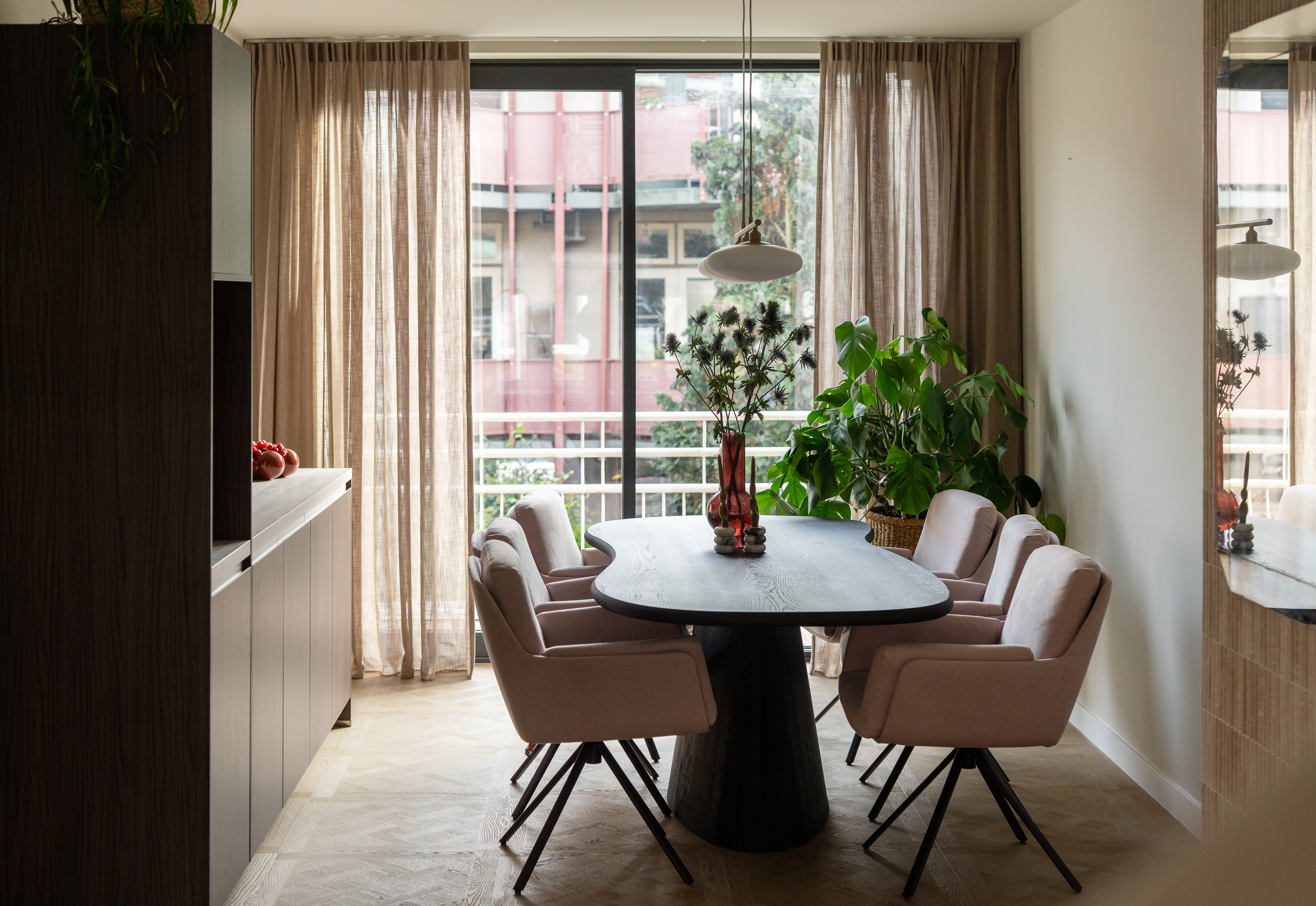 De afbeelding toont een moderne eetkamer met een grote schuifpui die natuurlijk licht binnenlaat. De pui is omgeven door lichte gordijnen, wat een zachte, luchtige sfeer creëert. Buiten is een balkon met een reling en wat groen te zien.