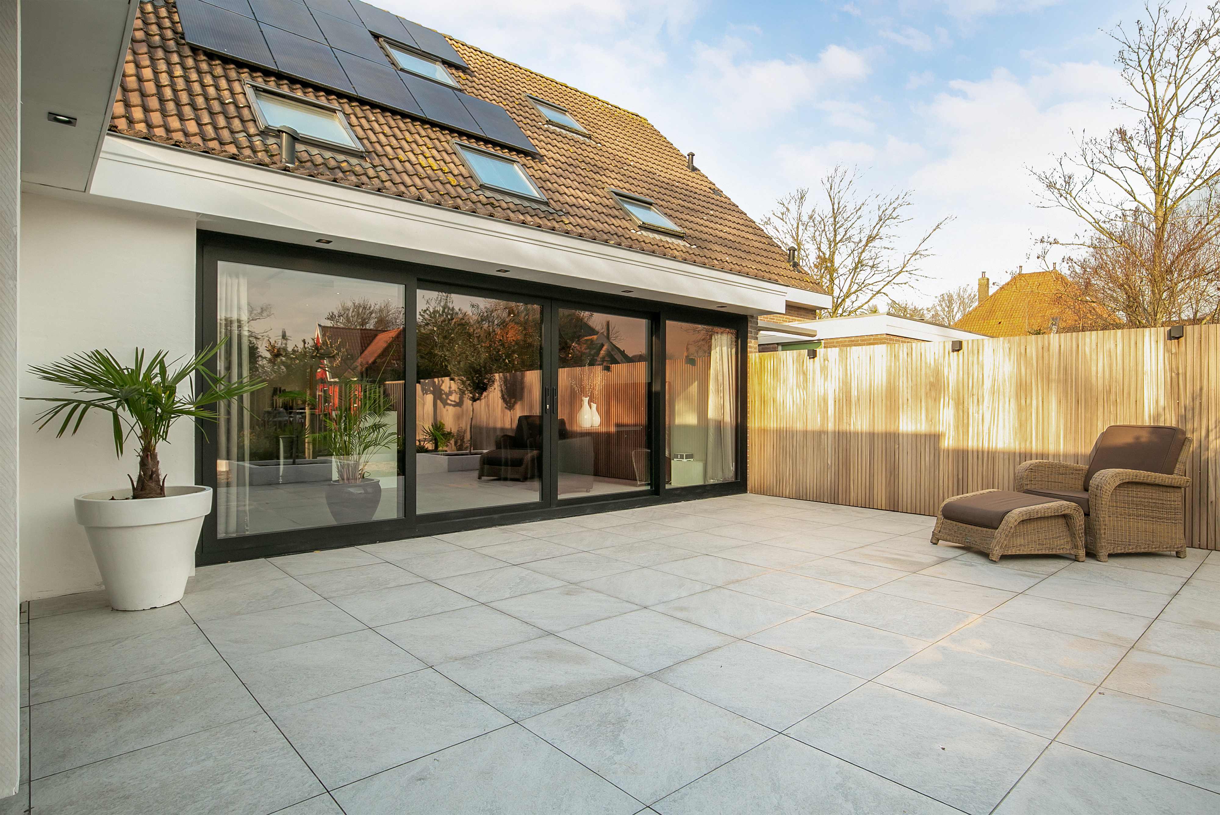 De afbeelding toont een modern huis met grote schuifpuien die uitkomen op een ruime patio. De kozijnen zijn zwart, wat een strak contrast geeft met de witte muren. Op het dak zijn er verschillende dakramen die natuurlijk licht binnenlaten. De patio is betegeld en heeft buitenzitplaatsen, wat een naadloze binnen-buiten leefruimte creëert.