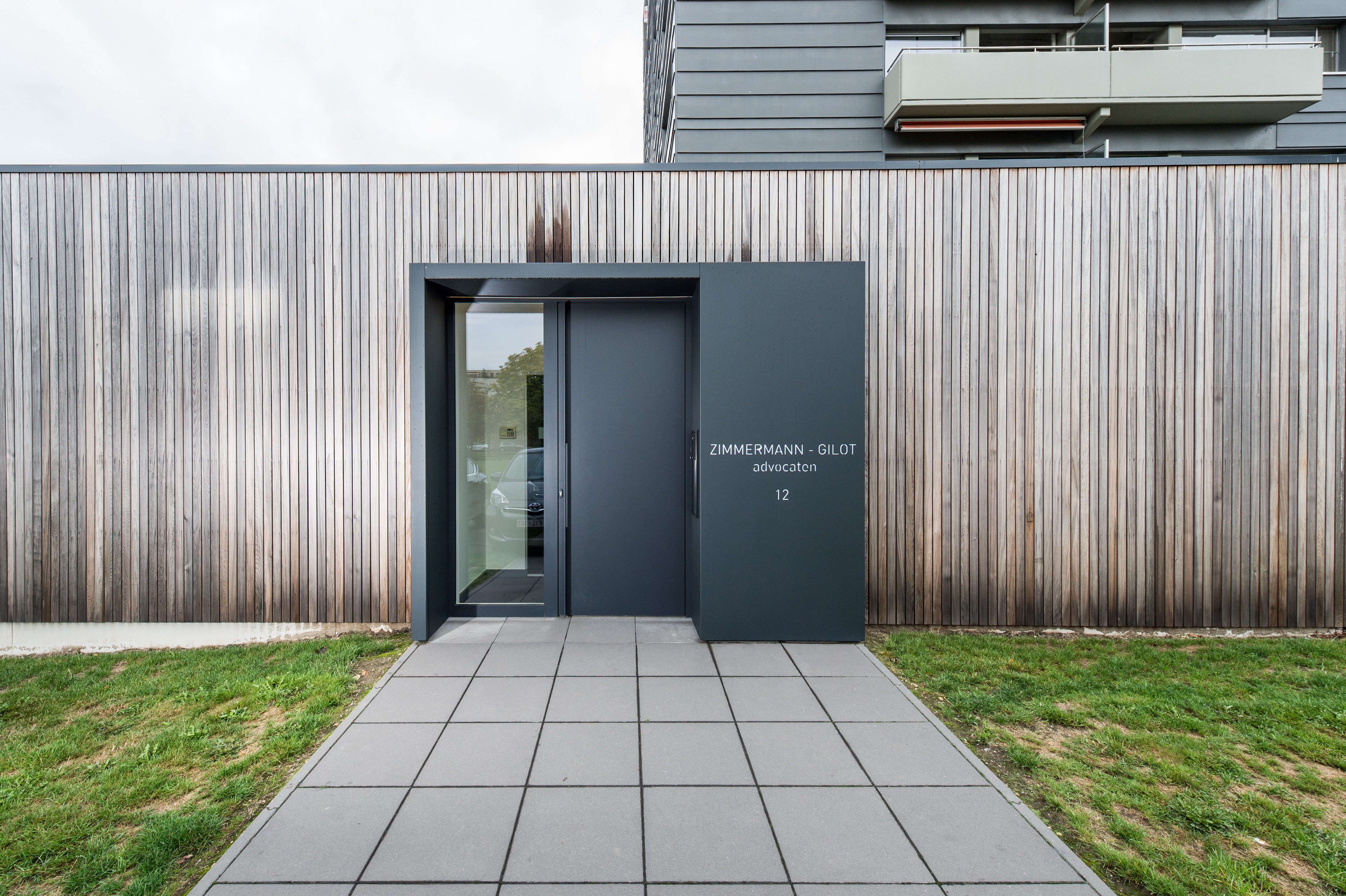 L'image montre une façade moderne avec une porte sombre et élégante et un grand châssis. La porte est intégrée dans un mur en bois, offrant un look minimaliste et contemporain. Le châssis à côté de la porte permet à la lumière naturelle d'entrer, améliorant l'esthétique du bâtiment.