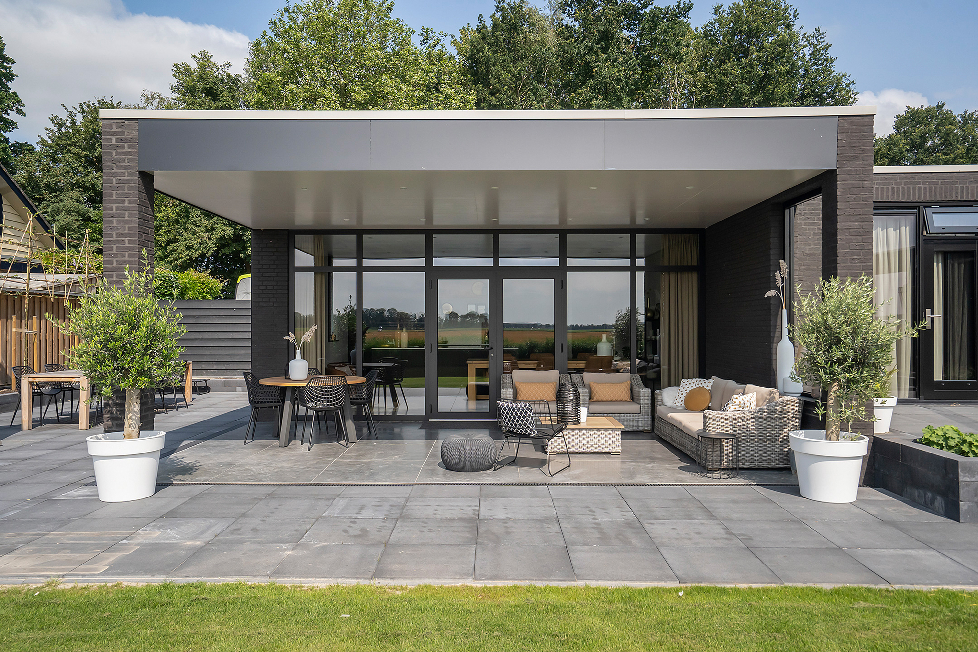 Het moderne huis heeft grote schuifpuien en deuren die uitkomen op een ruime patio. De glazen panelen zorgen voor een naadloze overgang tussen binnen en buiten, waardoor natuurlijk licht het interieur binnenstroomt. Het strakke ontwerp vult de eigentijdse architectuur aan en verhoogt de esthetische aantrekkingskracht van de woning.