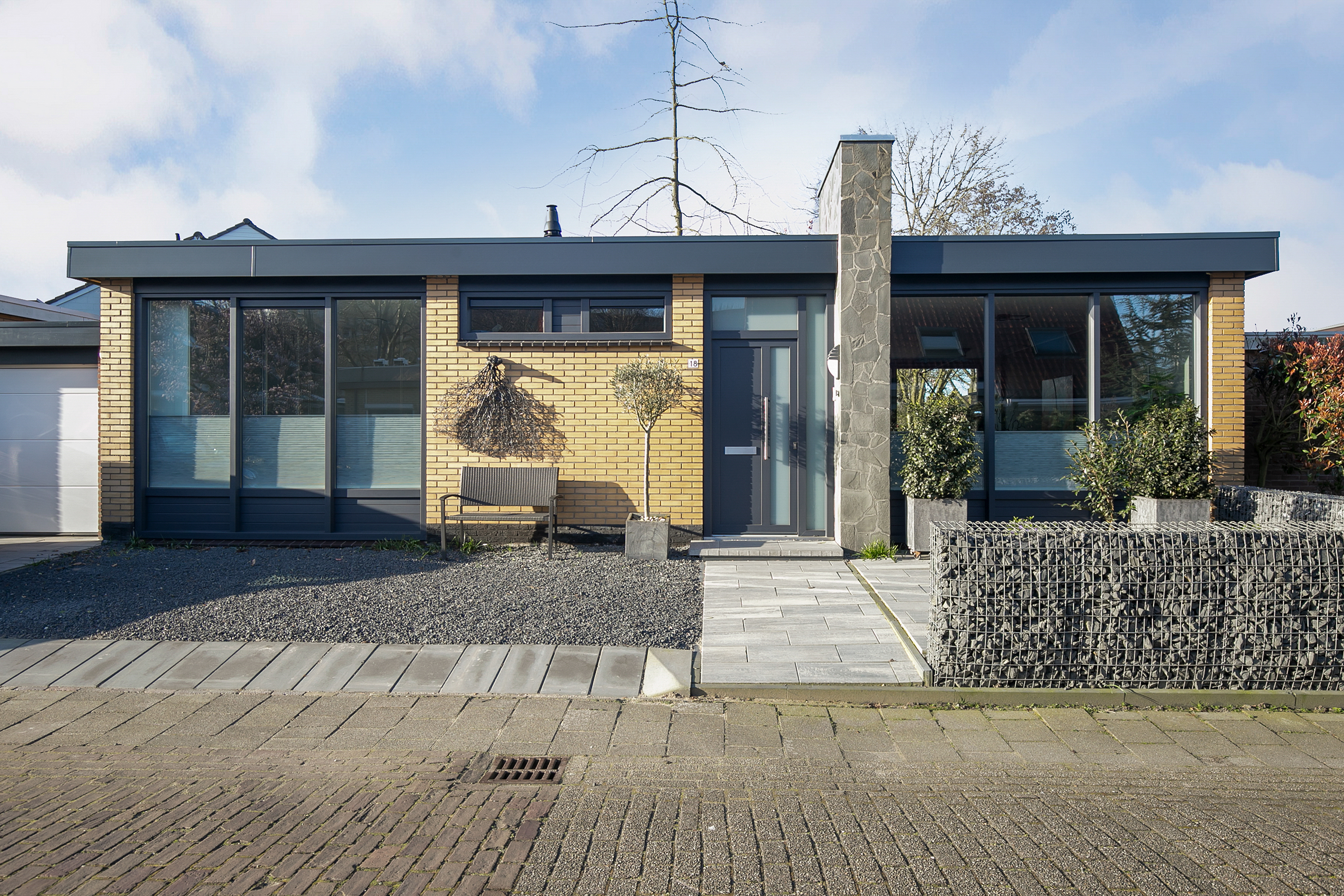 Het moderne huis heeft grote schuifpuien en een strakke voordeur. De kozijnen laten veel natuurlijk licht binnen, wat het eigentijdse ontwerp van het huis versterkt. De deur vult de kozijnen aan met zijn minimalistische stijl.