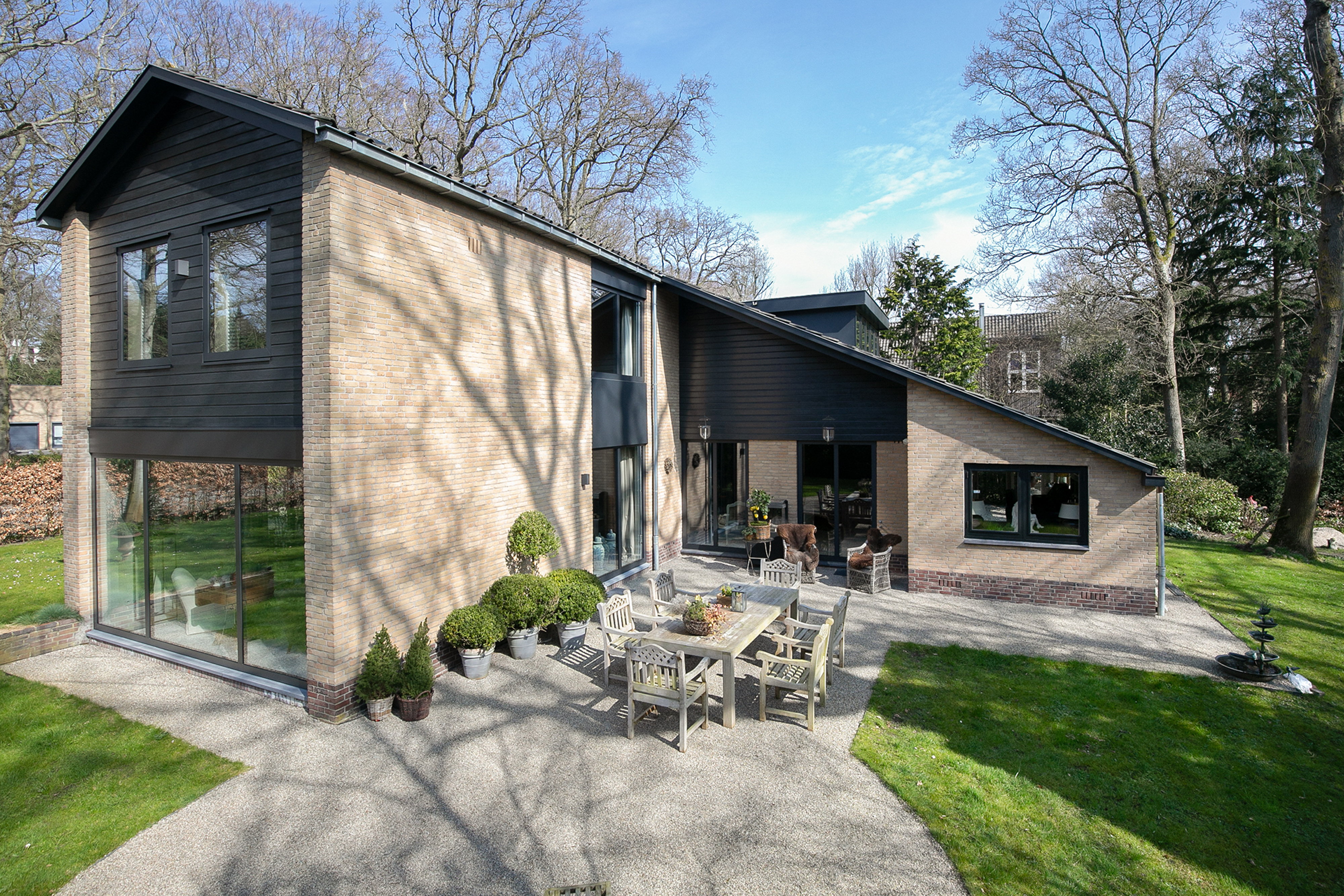 Het moderne huis heeft grote schuifpuien en deuren die uitkomen op een ruim terras. De kozijnen zijn strak zwart, wat mooi past bij de bakstenen gevel. De schuifpuien bieden gemakkelijke toegang tot de buitenzitplaats, wat de binnen-buitenstroom bevordert.
