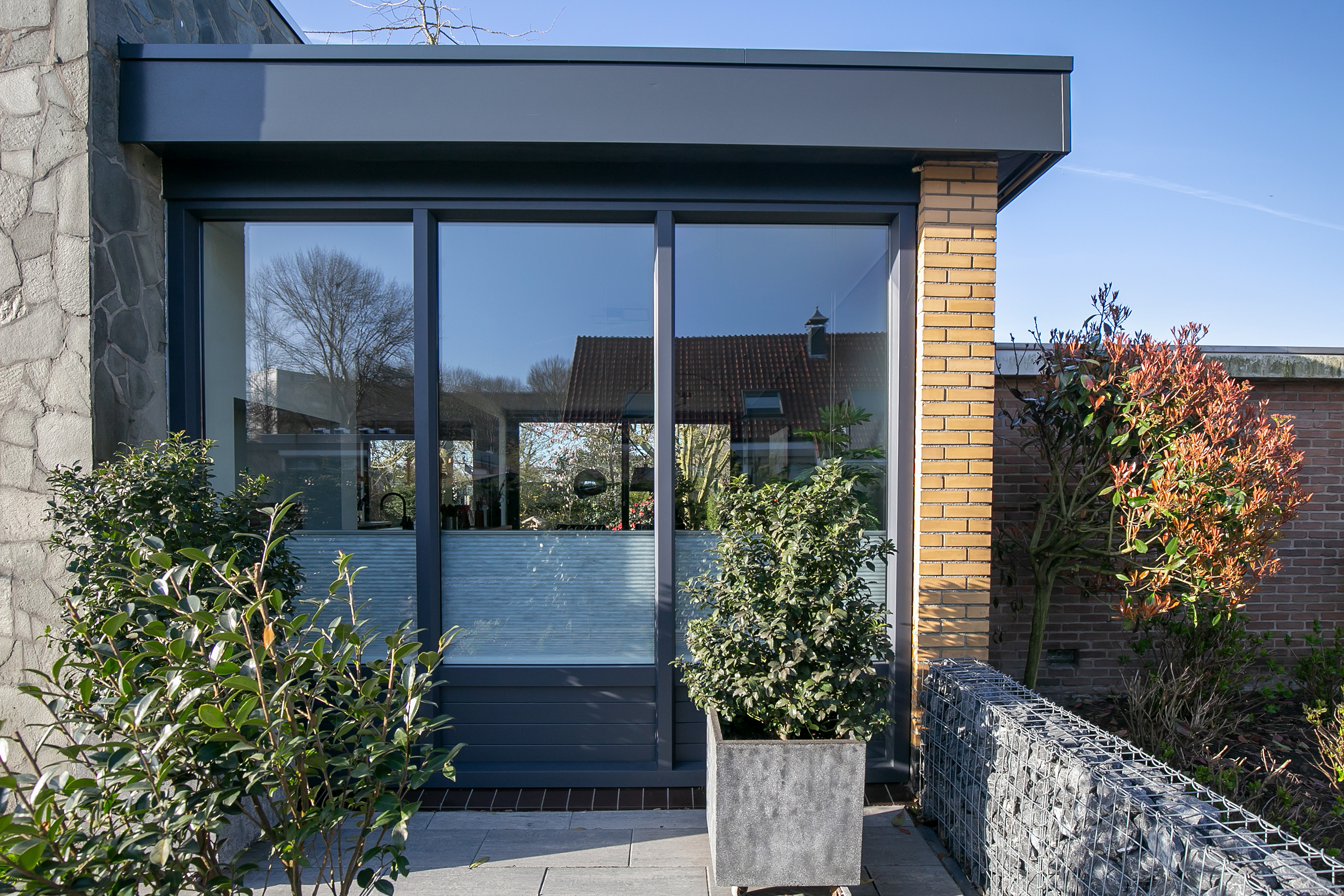 De afbeelding toont een moderne huisgevel met grote schuifpuien. De kozijnen zijn van donker metaal, wat een strakke en eigentijdse uitstraling geeft. Ze laten veel natuurlijk licht binnen. De omgeving heeft een mix van stenen en bakstenen muren, wat textuur en contrast aan het ontwerp toevoegt.