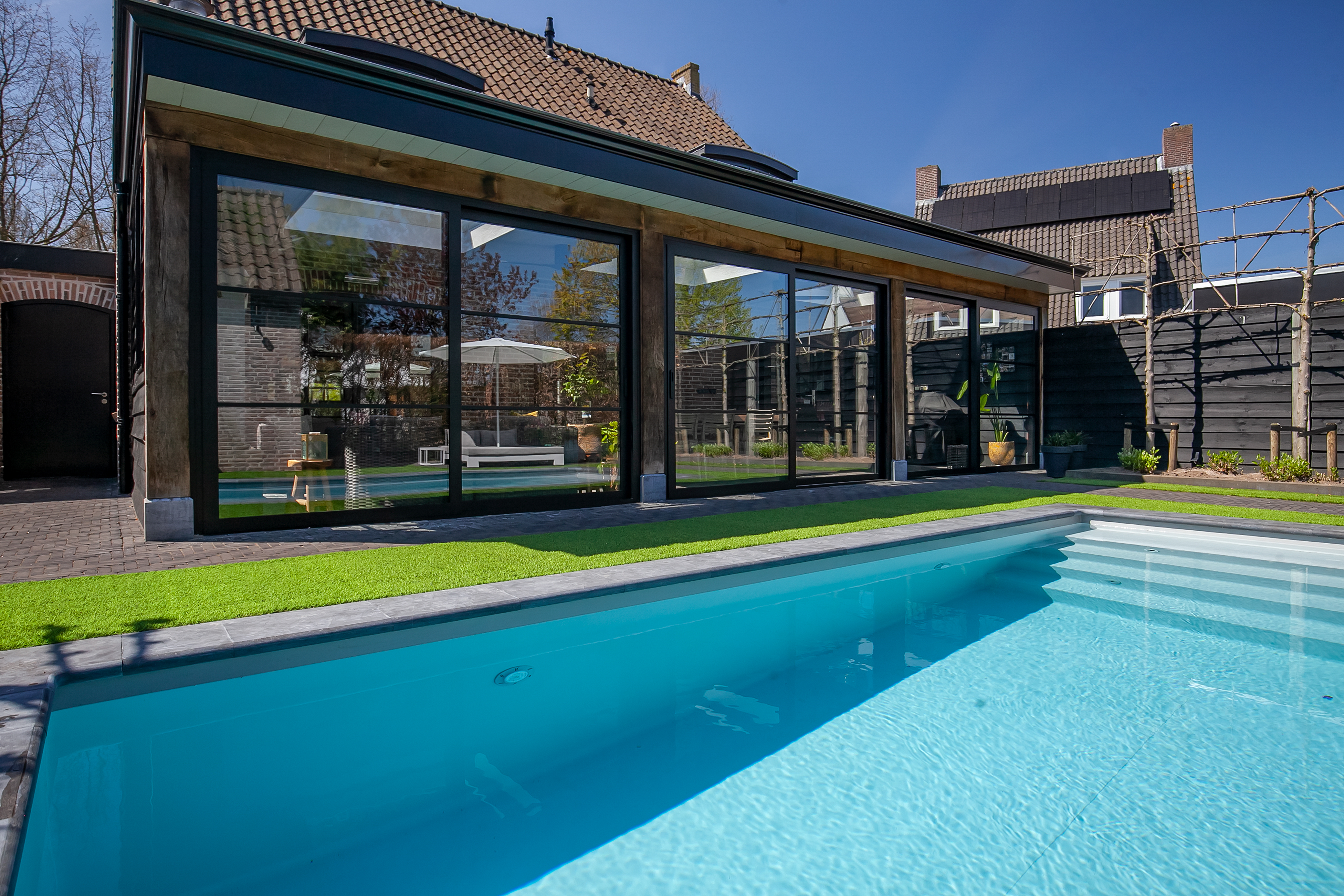 De afbeelding toont een modern huis met grote schuifpuien die uitkijken op een tuin met een zwembad. De kozijnen zijn van donker metaal, wat een strakke en eigentijdse uitstraling geeft. De deuren zijn geïntegreerd in het ontwerp van de kozijnen, wat zorgt voor een naadloze overgang tussen binnen en buiten. De weerspiegeling van de tuin en het zwembad in het glas verhoogt de esthetische aantrekkingskracht.