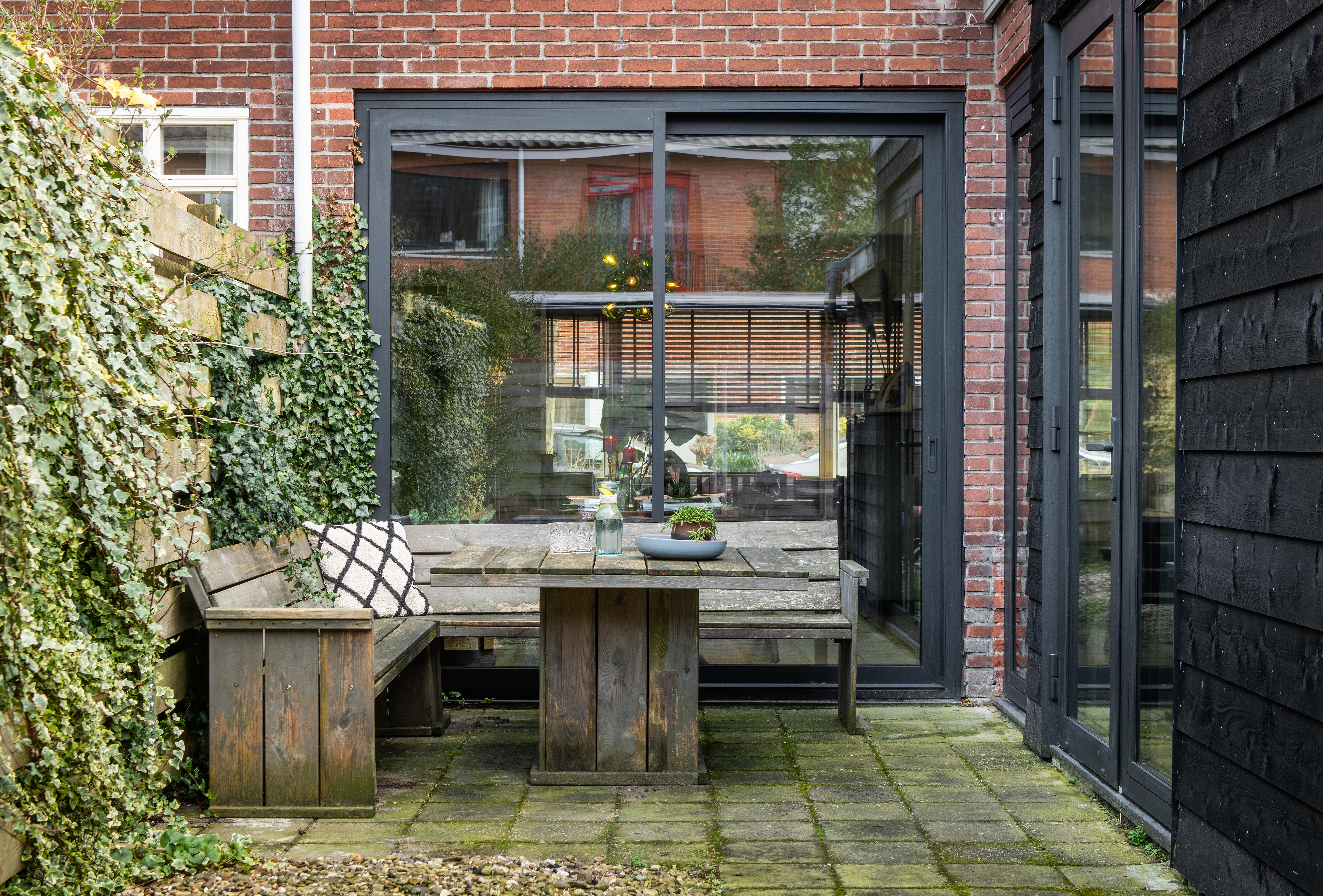 De afbeelding toont een gezellige buitenpatio met grote schuifpuien en deuren. De schuifpuien hebben een donker metalen frame, wat een modern contrast biedt met de rode bakstenen muren. De deuren openen naar een kleine tuin, wat de binnen-buitenstroom bevordert. De kozijnen weerspiegelen het groen buiten, wat bijdraagt aan de serene sfeer.