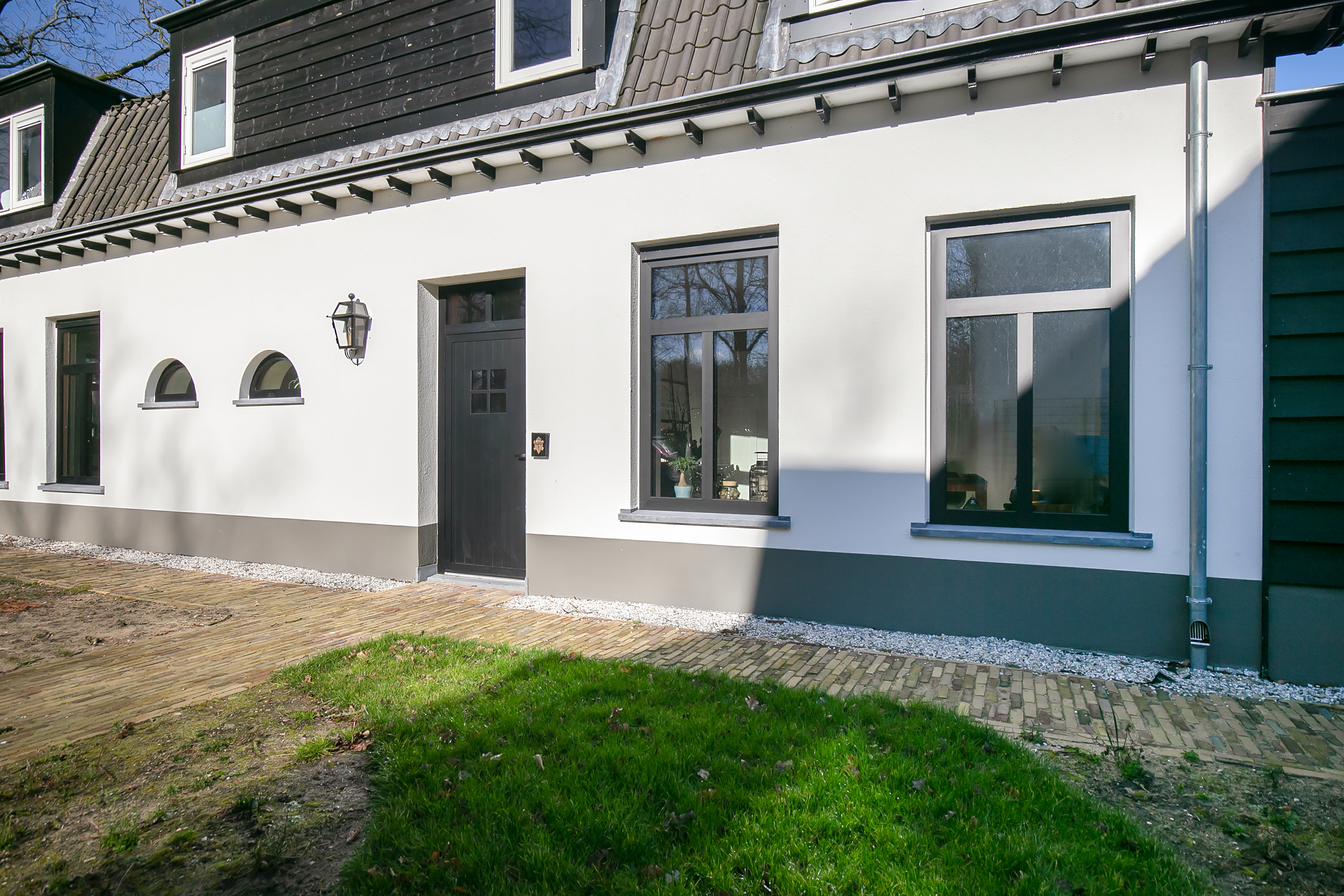 De afbeelding toont een moderne huisgevel met een combinatie van rechthoekige en gebogen kozijnen. De rechthoekige kozijnen zijn groot en laten veel licht binnen. De gebogen kozijnen voegen een uniek architecturaal element toe. De deur is centraal geplaatst en donker geschilderd, wat mooi past bij de kozijnen.