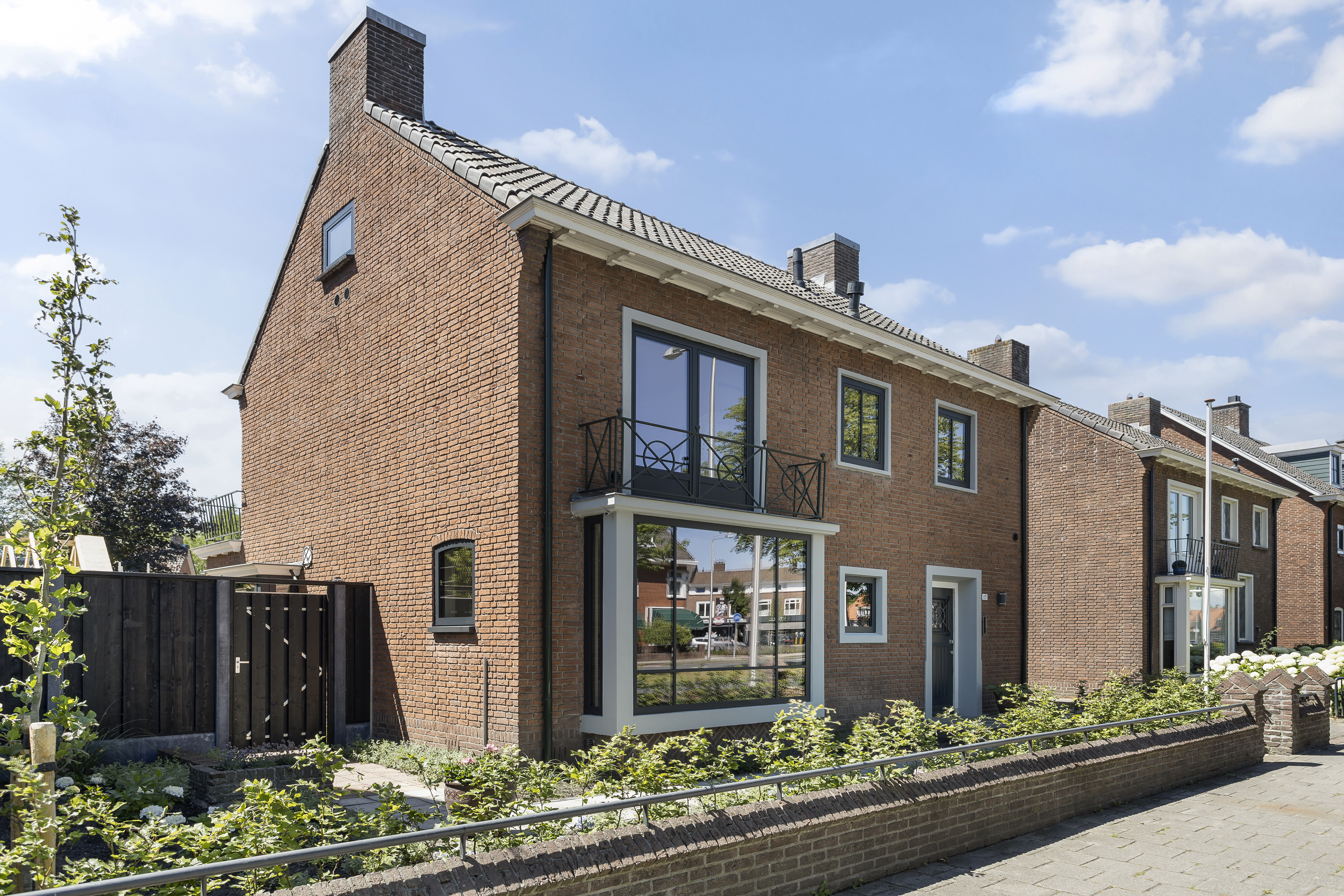 De afbeelding toont een bakstenen huis met verschillende kozijnen en een deur. Aan de voorkant is er een groot erkerkozijn en een schuifpui die leidt naar een klein balkon. De kozijnen zijn wit omlijst, wat contrasteert met de rode baksteen. Een houten deur is zichtbaar, passend bij de traditionele stijl van het huis.